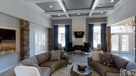 This thoughtfully designed single-floor space makes a strong architectural impression from the moment you enter. The centerpiece of the living area is a dramatic coffered ceiling with recessed lighting, painted in deep charcoal tones and framed by crisp white molding, lending a sense of grandeur and refined sophistication. Tall windows dressed in rich navy curtains flood the space with natural light while offering a polished, formal aesthetic.

A sleek, floor-to-ceiling white fireplace serves as a bold focal point, flanked by symmetrically arranged dark accent chairs that create an intimate seating arrangement. The herringbone-patterned hardwood flooring adds a classic yet contemporary texture underfoot. Complementing this are plush sofas in warm taupe tones, layered over a shaggy area rug, striking a balance between comfort and elegance.

Decorative Buddha sculptures positioned near the windows introduce a serene, curated quality to the room. A wall-mounted flat-screen television above the fireplace seamlessly integrates modern functionality into the refined setting. French doors on one side provide additional architectural character and natural light. The overall ambiance is one of luxurious calm, blending classic design elements with modern touches to create a welcoming yet visually striking space.