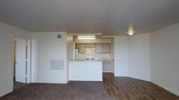 This single-floor residence showcases an open-concept design that seamlessly connects the kitchen and living areas. The kitchen is positioned at the center of the space, framed by a half-wall pass-through counter that allows for easy interaction between cooking and living areas. It is equipped with light wood cabinetry, a white refrigerator, a white range, and an under-cabinet range hood, all illuminated by a fluorescent overhead light fixture. The cabinetry provides ample storage, and the layout supports functional meal preparation. The living area features a notable transition in flooring, with plush carpet occupying one section and wood-style hard flooring in another, adding visual interest and defined zones within the open plan. The walls are painted in a neutral white tone, contributing to a clean and airy atmosphere. Ventilation is supported by visible ceiling and wall-mounted HVAC vents throughout the space. Recessed and surface-mounted ceiling light fixtures provide consistent artificial lighting across the unit. The overall layout is practical and straightforward, making efficient use of the available space across its single level.