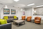 The Media Center is a thoughtfully designed single-floor communal space that offers a comfortable and inviting environment for both entertainment and relaxed social gatherings. The layout is divided into two connected lounge areas, each serving a distinct purpose while maintaining a cohesive aesthetic throughout.

The primary lounge area is anchored by a large flat-screen television mounted on a modern media console, making it ideal for group viewing. Plush charcoal leather sofas accented with vibrant yellow-green throw pillows provide ample seating, complemented by a sleek rectangular coffee table. Natural light filters in through windows dressed in olive-green grommet curtains, adding warmth to the neutral gray walls and patterned carpeted flooring. Crown molding and recessed panel lighting add refined architectural detail to the space.

The secondary area features a cluster of tan leather swivel chairs arranged around small side tables, offering a more conversational and flexible seating arrangement. A built-in counter with white cabinetry and a wood-plank accent wall serves as a stylish backdrop, ideal for refreshments or décor display. Coordinating arc floor lamps and framed artwork are distributed throughout both areas, contributing to a polished and cohesive interior design. Together, the two zones create a versatile and welcoming Media Center suited for a variety of group activities.