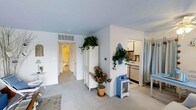 This single-floor space presents a clean, open layout characterized by white walls and a distinctive soft blue ceiling that lends a calm, airy quality to the interior. The main living area features neutral carpeting and flows naturally toward a compact kitchen zone, partially visible and equipped with standard appliances including a refrigerator. A white wardrobe cabinet serves as a functional room divider, while trailing green plants mounted on walls and atop furniture introduce a fresh, botanical element throughout. A small blue accent cabinet and a potted flowering plant add pops of color against the predominantly white palette. A hallway leads toward a bathroom, visible in the background with warm lighting, suggesting a well-connected floor plan. Curtained partitions on one side hint at additional functional zones within the space. The overall design balances simplicity with decorative charm, using natural elements and pastel tones to create a welcoming, cohesive environment suited for comfortable everyday living.