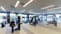 This contemporary fitness center is designed to deliver a premium workout experience within a clean, open, and well-organized space. The facility is laid out on a single floor, allowing for an intuitive flow between different exercise zones. Large floor-to-ceiling windows line the perimeter, flooding the space with natural light and providing open views of the surrounding outdoor greenery, creating an energizing and refreshing atmosphere for exercise.

The cardio zone is prominently featured and includes multiple treadmills and elliptical machines arranged to take full advantage of the scenic window views. Alongside these, a well-curated selection of strength and resistance training machines is positioned throughout the space, offering users a comprehensive range of workout options. Additional free-weight and functional training areas are also visible, catering to a variety of fitness routines.

The interior finishes are sleek and modern, with a neutral-toned rubberized floor that provides both safety and comfort underfoot. Overhead, linear LED lighting runs across the white ceiling, ensuring bright, even illumination throughout the entire space. The overall ambiance is clean, professional, and inviting — reflecting a facility built with both performance and comfort in mind.