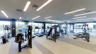 This contemporary fitness center is designed to deliver a premium workout experience within a clean, open, and well-organized space. The facility is laid out on a single floor, allowing for an intuitive flow between different exercise zones. Large floor-to-ceiling windows line the perimeter, flooding the space with natural light and providing open views of the surrounding outdoor greenery, creating an energizing and refreshing atmosphere for exercise.

The cardio zone is prominently featured and includes multiple treadmills and elliptical machines arranged to take full advantage of the scenic window views. Alongside these, a well-curated selection of strength and resistance training machines is positioned throughout the space, offering users a comprehensive range of workout options. Additional free-weight and functional training areas are also visible, catering to a variety of fitness routines.

The interior finishes are sleek and modern, with a neutral-toned rubberized floor that provides both safety and comfort underfoot. Overhead, linear LED lighting runs across the white ceiling, ensuring bright, even illumination throughout the entire space. The overall ambiance is clean, professional, and inviting — reflecting a facility built with both performance and comfort in mind.