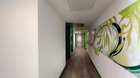 Located at 300 18th St S in Birmingham, Alabama, this residential corridor floor presents a striking and thoughtfully designed interior that makes a strong visual impression. The hallway system follows an angular layout, connecting multiple numbered units along its length, as revealed by the overhead floor plan view.

The design palette is defined by a dynamic use of green — ranging from deep forest tones to bright lime hues — applied to door frames, ceiling recesses, and baseboards. This layering of green shades creates a sense of depth and energy throughout the corridor. The white walls serve as a clean contrast, allowing the color accents to stand out boldly.

Large-scale botanical and nature-inspired murals are prominently displayed along the corridor walls, adding an artistic, organic quality to the space. These illustrated works feature flowing leaves and plant forms rendered in soft greens and grays, contributing to a calming yet vibrant atmosphere.

The flooring consists of durable, large-format gray tile laid in an offset pattern, providing a neutral grounding element that complements the vivid wall treatments. Recessed ceiling lighting ensures even illumination throughout the corridor, while subtle cove lighting near the green ceiling accents adds warmth and dimension. Room number plaques with backlit details are mounted beside each unit entry, offering both functional wayfinding and a polished aesthetic finish.