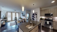 This well-appointed single-floor residence showcases a cohesive contemporary design with dark hardwood-style flooring, neutral tones, and modern fixtures throughout the main living areas. The open-concept kitchen serves as the heart of the home, anchored by a central island with a dark countertop, an undermount stainless steel sink, and pendant lighting overhead. Wood-toned cabinetry with sleek hardware lines the perimeter walls, complemented by a white subway tile backsplash and a full suite of stainless steel appliances, including a refrigerator, range, microwave, and dishwasher.

The kitchen flows seamlessly into the living and dining areas, where a ceiling fan and large windows with slatted blinds allow natural light to fill the space. A set of glass-paneled doors in the living area provides an additional connection to the outdoors.

The primary bedroom features plush carpeting, generous windows, a ceiling fan, and direct access to a private bathroom with a walk-in glass shower, a wood-toned double vanity with white countertops, and an adjoining walk-in closet with multi-level shelving. A second bedroom offers carpeted flooring and ample wall space. The second bathroom includes a tub with shower, a floating wood vanity, and light tile flooring.