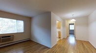 This single-floor space offers a clean, functional layout with a warm and inviting ambiance throughout. The interior is finished with smooth hardwood flooring that runs seamlessly across the living areas, catching natural light and adding a sense of warmth to the space. Crisp white walls and white trim provide a neutral backdrop that enhances the brightness of each room.

The main living area is open and airy, with a window that draws in natural light and a baseboard heating system integrated neatly along the walls. A partial wall creates a natural division between the living space and the kitchen, which is accessible through a wide open passageway. The kitchen features light wood cabinetry, providing ample storage, and is well-lit with a functional layout suited for everyday use.

A hallway extends toward the entry of the space, where a pendant light fixture hangs overhead, casting a warm glow. The entryway is fitted with a solid door that includes a window panel, allowing additional natural light to filter into the corridor and creating a welcoming first impression. The overall design is straightforward and practical, with consistent finishes and a layout that promotes easy flow between areas.