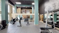Located at 712 Tucker St in Raleigh, NC, this fitness facility offers a spacious, thoughtfully designed workout environment spread across a single floor with a striking double-height ceiling that lends the space an open, airy feel. Teal-painted structural columns punctuate the floor plan, complementing the dark ceiling and creating a modern, energetic aesthetic. An upper mezzanine level overlooks the main workout floor below, connected by a railed walkway and providing additional equipment space visible from the ground level. The main floor is organized into distinct training zones. A cardio area lines a windowed wall, where large, tall windows flood the space with natural light and offer views of surrounding greenery, housing a row of treadmills and an elliptical machine. A dedicated free weights zone features a full dumbbell rack, adjustable benches, and step platforms arranged on durable rubber flooring. A strength training area showcases a multi-functional cable machine system and weight benches set against an exposed brick accent wall bearing the "Raleigh Office Supply" branding — a nod to the building's heritage. A decorative mural adorns one dark feature wall, adding artistic character. Private rooms with glass-door entries are also present, offering enclosed spaces for additional fitness activities. The combination of industrial-chic design elements, abundant natural light, and comprehensive equipment makes this a versatile and inviting fitness destination.