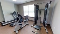 This single-floor fitness center is thoughtfully laid out to maximize functionality within a focused space. The room features a neutral color palette with light gray walls and durable speckled flooring well-suited to high-traffic exercise use. Natural light filters in through a large window fitted with horizontal blinds, creating a bright and energizing atmosphere for workouts.

The cardio section is outfitted with multiple machines, including an elliptical trainer and a recumbent stationary bike, catering to a range of low-impact cardiovascular exercise preferences. For strength training, the space includes a multi-function cable machine complete with a pull-down bar and an adjustable workout bench, offering a variety of resistance exercises targeting different muscle groups. Additional strength equipment is visible along the walls, further expanding training options.

The layout is practical and efficient, with equipment arranged to allow adequate movement between machines. Ventilation is supported by ceiling vents, contributing to a comfortable environment during use. Overall, this fitness center provides a clean, functional, and well-maintained space with the essential equipment needed to support both cardio and strength-based fitness routines.