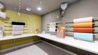 This purpose-built gift wrapping room is thoughtfully designed to provide a fully equipped, organized space for all gift preparation needs. The room features a generous L-shaped white countertop that wraps along two walls, offering ample workspace for wrapping, assembling, and decorating gifts of all sizes. Mounted wall rails line multiple walls, providing convenient and accessible storage for rolls of wrapping paper, keeping supplies neat and within easy reach. The interior design balances function with a cheerful aesthetic — one wall is painted in a bold mustard yellow, while the remaining walls are finished in a warm grey tone, creating an energetic yet cohesive atmosphere. Recessed lighting illuminates the workspace evenly from above, while a ceiling ventilation system ensures comfortable airflow throughout. The neutral grey flooring grounds the space and complements the overall color palette. The room is well-suited for both personal and communal use, making it an ideal amenity within a larger residential or retail facility. Its compact yet efficient layout ensures that everything a gift-giver needs — from wrapping paper and gift bags to ribbon and tissue — is organized, accessible, and ready to use.