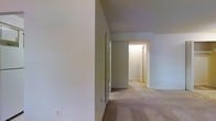 This single-floor space is characterized by a straightforward, functional layout centered around a carpeted hallway that serves as the main circulation path connecting the various rooms. The flooring throughout consists of wall-to-wall carpet in a neutral beige tone, contributing to a quiet and cohesive feel underfoot. The walls are finished in a soft, light gray-white palette that keeps the interior feeling clean and understated. The hallway extends toward what appears to be additional rooms, with doorways visible along its path, including a louvered door that likely conceals a closet or utility space, providing practical storage. Overhead lighting illuminates the corridor, ensuring the space remains well-lit despite the absence of visible windows in this area. Electrical outlets are positioned at standard heights along the baseboards, and a floor vent is visible, indicating a central HVAC system. The overall ambiance is calm and neutral, offering a blank-canvas quality that allows for flexible use and personalization. The space presents a no-frills, practical interior that prioritizes functionality and ease of maintenance.