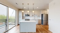 This compact, single-floor unit offers a thoughtfully designed layout that maximizes functionality within its efficient footprint. The kitchen serves as the centerpiece of the living space, showcasing white shaker-style cabinetry, a diamond-pattern tile backsplash, and light quartz countertops. A central island provides additional prep and seating space, illuminated by three globe pendant lights suspended from the ceiling on brass rods. Stainless steel appliances, including a range, microwave, dishwasher, and refrigerator, complete the well-equipped kitchen. Light hardwood flooring runs throughout the main living area, enhancing the bright, airy atmosphere. A large sliding glass door opens to a private balcony, allowing natural light to flood the space and offering outdoor views. A hallway lined with barn-style sliding doors connects the main living area to the bedroom and bathroom. The bedroom features neutral carpet and clean white walls, providing a calm retreat. The bathroom includes a full soaking tub with subway-tiled surround and polished chrome fixtures, maintaining the unit's clean, modern aesthetic throughout.