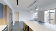 This thoughtfully designed single-floor space showcases a seamless open-concept layout that connects the kitchen and living areas into one cohesive and functional environment. The kitchen is anchored by a generous island with a sleek white quartz countertop and warm wood cabinetry below, creating an inviting contrast of tones and materials. A striking row of modern pendant lights hangs above the island, providing both focused task lighting and a stylish design focal point. Stainless steel appliances, including a built-in wine cooler, add a refined, contemporary touch. The floors throughout are rich dark hardwood, grounding the space with warmth and texture against the crisp white walls and ceilings. Recessed lighting and track lighting supplement the natural light that filters in through a window overlooking the surrounding urban landscape. The living area extends naturally from the kitchen, offering an open and airy feel well-suited for both relaxing and entertaining. The overall aesthetic is clean, minimal, and modern, with a neutral palette that provides a versatile backdrop for a variety of interior styles. The space is move-in ready and presents a polished, well-appointed environment from every angle.