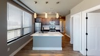 This single-floor residence offers a thoughtfully designed layout that balances modern style with everyday functionality. The open-concept kitchen serves as the heart of the space, anchored by a large central island with a built-in sink and crisp white countertops. Rich, dark cabinetry lines the walls and is complemented by a classic white subway tile backsplash and a full suite of stainless steel appliances, including a refrigerator, electric range, and over-the-range microwave. Warm wood-tone flooring runs throughout the kitchen and main areas, while pendant lighting and a track light system above provide both task and ambient illumination. Natural light enters generously through multiple windows fitted with horizontal blinds. The two bedrooms are carpeted for comfort and each feature a ceiling fan with lighting and a window overlooking the exterior. The primary bathroom includes a bathtub and shower combination, wood-look flooring, and a louvered storage closet. The second bathroom is finished with a dark wood vanity, a white countertop, a large mirror, and a sleek backlit light fixture with matte black hardware accents throughout. The overall aesthetic is clean, contemporary, and move-in ready.