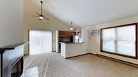 This single-floor space showcases an open-concept design that connects the living area and kitchen in a fluid, functional layout. The living area is finished with plush wall-to-wall carpeting and benefits from abundant natural light streaming through large sliding glass doors fitted with vertical blinds, creating a bright and welcoming atmosphere. A ceiling fan with a light fixture is mounted to the vaulted ceiling, adding both airflow and a sense of vertical spaciousness overhead.

The kitchen is positioned toward the rear of the open layout and features warm-toned wood cabinetry, dark countertops, and a peninsula that serves as a natural divider between cooking and living spaces. Stainless steel appliances, including a refrigerator, range, and microwave, are integrated into the design, providing a modern and practical cooking environment. A pendant light hangs above the peninsula, adding focused illumination to the workspace.

A wall-mounted air conditioning unit and a baseboard heater offer dedicated climate control throughout the space. A window on the far wall allows additional natural light to filter in. Overall, the space presents a clean, functional, and well-lit environment.