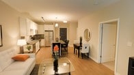 This single-floor residence showcases an open-concept design that seamlessly connects the kitchen, dining, and living areas, creating a cohesive and functional living environment. The kitchen is outfitted with stainless steel appliances, including a full-size refrigerator and range, complemented by white cabinetry and a dark tile backsplash that adds visual contrast. A peninsula with bar seating provides a casual dining option, while pendant lighting above enhances the space with a warm, inviting glow. Adjacent to the kitchen, a dedicated dining area accommodates a dark-toned table with upholstered white chairs, accented by a decorative plant and a round wall mirror that adds depth to the room. The living area, positioned at the front of the space, features a glass coffee table and a patterned area rug over light hardwood flooring that runs throughout the unit. Warm recessed and track lighting creates an intimate ambiance across all areas. A dark-painted entry door anchors the far end of the space, with additional rooms accessible through doorways along the right wall, suggesting private sleeping or bathroom quarters beyond the open living zone.