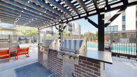 Located at 18 N Orange St in Salt Lake City, Utah, this single-level outdoor amenity space is designed for communal gathering and leisure. The centerpiece of the area is a substantial wood-and-steel pergola that spans the length of the patio, providing partial shade while allowing natural light to filter through its open lattice framework. Strands of globe lights are draped throughout the pergola, creating an inviting ambiance for both daytime and evening use.

Beneath the pergola, a built-in outdoor kitchen features two large stainless steel gas grills set into a brick-clad counter with a dark stone work surface, offering a functional setup for outdoor cooking. Adjacent to the grill station, a seating area is arranged around a gas fire pit with blue glass media, furnished with a mix of woven lounge chairs, rocking chairs, and a loveseat with cushions in bold accent colors.

The patio connects directly to a gated swimming pool area enclosed by wrought iron fencing, with mature trees and landscaping providing a natural backdrop. The surrounding multi-story residential building frames the space, while the open-air design ensures ample airflow and sunlight throughout the day.