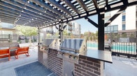 Located at 18 N Orange St in Salt Lake City, Utah, this single-level outdoor amenity space is designed for communal gathering and leisure. The centerpiece of the area is a substantial wood-and-steel pergola that spans the length of the patio, providing partial shade while allowing natural light to filter through its open lattice framework. Strands of globe lights are draped throughout the pergola, creating an inviting ambiance for both daytime and evening use.

Beneath the pergola, a built-in outdoor kitchen features two large stainless steel gas grills set into a brick-clad counter with a dark stone work surface, offering a functional setup for outdoor cooking. Adjacent to the grill station, a seating area is arranged around a gas fire pit with blue glass media, furnished with a mix of woven lounge chairs, rocking chairs, and a loveseat with cushions in bold accent colors.

The patio connects directly to a gated swimming pool area enclosed by wrought iron fencing, with mature trees and landscaping providing a natural backdrop. The surrounding multi-story residential building frames the space, while the open-air design ensures ample airflow and sunlight throughout the day.