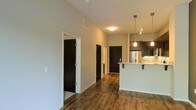 This single-floor space is defined by a thoughtful open-concept design that connects the main living area to a well-appointed kitchen. Rich, dark hardwood flooring runs throughout, adding warmth and continuity to the layout. The elevated ceilings contribute a sense of spaciousness, allowing light to travel freely across the neutral-toned walls.

The kitchen stands out as a focal point, featuring a peninsula with bar seating, pendant lighting suspended from above, and track lighting to accent the workspace. Dark cabinetry pairs with a tile backsplash in warm tones, while a stainless steel refrigerator anchors the back wall. The countertops appear to be a light-colored stone or composite, providing a striking contrast to the darker elements.

A central hallway leads to multiple rooms, each accessed through dark-finished doors that complement the flooring. The consistent design language throughout — clean white walls, dark wood accents, and modern fixtures — creates a cohesive and contemporary ambiance. The overall layout feels functional and well-organized, balancing open communal space with clearly defined private areas.