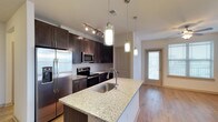 This 2-bedroom, 2-bathroom single-floor residence offers a well-designed layout with modern finishes throughout. The kitchen serves as a central highlight, featuring rich dark wood cabinetry that contrasts elegantly with light granite countertops on the island, which includes an undermount sink and a sleek gooseneck faucet. A suite of stainless steel appliances — including a French door refrigerator with ice and water dispenser, a built-in microwave, and an electric range — provides both functionality and a cohesive aesthetic. Overhead, a combination of track lighting and cylindrical pendant lights illuminate the space effectively, adding a contemporary touch. The kitchen flows naturally toward the entryway, where a full-lite door allows natural light to filter into the space, brightening the warm hardwood-style flooring that runs throughout the main living areas. The overall design balances practicality with modern style, making the kitchen and adjoining spaces inviting and functional for everyday living.