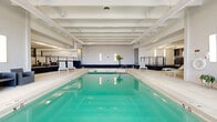 Located at 8000 E Girard Ave in Denver, Colorado, this Pool & Outdoor Living Space is a generously sized amenity facility spanning a substantial footprint across a single level. The centerpiece of the indoor area is a large lap pool with vivid turquoise water, surrounded by light-toned tile decking and complemented by white lounge chairs and modular seating. The pool room features a bold exposed beam ceiling finished in white, creating a clean, modern aesthetic, while vertical wall sconces provide even ambient lighting throughout. A mezzanine-level walkway with sleek black cable railings overlooks the pool, adding architectural depth and offering convenient access between areas. An adjacent lounge zone features rows of reclining chaise lounges positioned beside floor-to-ceiling glass walls and black-framed doors, which open directly to the outdoor living space. The outdoor patio is expansive, with smooth concrete flooring and a collection of wicker lounge furniture, armchairs, and bar-height seating with a grill station — ideal for social gatherings. A pergola structure provides partial shade, while mature trees and open sky complete the setting. The surrounding building facade, finished in bold orange and gray tones, frames the courtyard and adds vibrant architectural character to this well-appointed outdoor retreat.