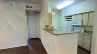 This single-floor residence offers a well-organized and functional layout designed for comfortable everyday living. The kitchen is a standout feature, designed with an open-concept style that incorporates a peninsula breakfast bar with a neutral countertop, providing both additional workspace and a casual dining option. White appliances, including a refrigerator and microwave, complement the bright, clean aesthetic of the kitchen. A built-in wine rack adds a charming and practical storage detail. The kitchen flows naturally into the hallway, which is finished with rich, dark hardwood flooring that extends throughout the space, adding warmth and visual continuity. Crown molding runs along the ceiling, lending a classic and refined character to the interiors. The ceilings are fitted with recessed ventilation and bright overhead lighting, ensuring the space feels well-lit and airy. White interior doors with panel detailing are present throughout, maintaining a cohesive and tidy design language. The overall ambiance is clean, bright, and inviting, with thoughtful architectural details that elevate the everyday functionality of the home into a polished and comfortable living environment.