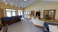 This single-story space offers a bright and welcoming interior characterized by soaring vaulted ceilings that lend an open, airy quality to the room. The walls are painted in a warm yellow tone, creating an inviting atmosphere complemented by neutral-toned carpet flooring throughout. Pendant lighting fixtures hang from the high ceiling, providing focused illumination alongside recessed lighting that brightens the space evenly.

A prominent bar-height seating area is positioned near large windows along one wall, allowing natural light to filter into the room. The striped high-back chairs arranged at the counter add a classic, refined touch. Warm wood-trimmed partitions divide the interior into functional zones without compromising the open feel of the layout.

Decorative elements, including sculptural bird figurines, a small potted plant, and an ornate dark wood cabinet with display items, add character and visual interest. A framed mirror on one wall enhances the sense of depth. French-style doors near the seating area connect the interior to the outside, further contributing to the space's light-filled, welcoming ambiance.