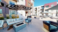 This expansive outdoor courtyard is a thoughtfully designed communal amenity space set within a modern multi-story residential community. The open-air layout is anchored by a generous concrete patio that connects multiple distinct seating zones, creating a versatile environment suited for both relaxed gatherings and lively socializing.

At the heart of the space are two lounge areas, each centered around a rectangular fire pit and furnished with plush, light-blue upholstered sofas and chairs. Large striped cantilever umbrellas provide shade overhead, while ornamental grasses in tall planters add a natural, decorative element. Additional seating clusters, featuring contemporary armchairs with wood-accented frames and gray cushions, are positioned throughout the courtyard under coordinating umbrellas.

A standout feature is the pergola-covered outdoor kitchen station, complete with a stainless steel built-in grill set into a handsome stacked-stone surround. The slatted pergola roof filters natural light while providing partial shelter, making this area ideal for outdoor cooking and entertaining.

The perimeter of the courtyard is framed by black metal railings, beyond which open views of the surrounding neighborhood and landscaping extend under a bright, clear sky. Warm brick and contemporary siding on the adjacent multi-story buildings lend architectural character to the setting, creating an inviting and well-appointed outdoor retreat.