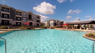 This outdoor pool area serves as a central amenity space designed for relaxation and recreation. The generously sized swimming pool features sparkling, crystal-clear turquoise water and is complemented by a separate spa or hot tub positioned nearby. A broad concrete pool deck surrounds the water, providing ample space for lounge chairs and seating arrangements shaded by vibrant umbrellas, offering residents comfortable spots to unwind under open skies.

Access to the pool area is provided through a dedicated pool house structure, characterized by stucco exterior walls, dark wood-framed doors, and wall-mounted lantern-style lighting. Posted pool rules signage is visible near the entrance, and potted planters add a touch of greenery to the entry area.

The pool is enclosed within a fenced perimeter, with mature trees and landscaping softening the boundaries of the space. Multi-story residential buildings with balconies surround the area, creating an enclosed, community-oriented atmosphere. Large glass sliding doors from adjacent units offer direct sightlines to the pool, enhancing the sense of connectivity between interior living spaces and the outdoor amenity. The open-sky setting allows abundant natural light throughout the day, contributing to a bright and inviting outdoor environment.