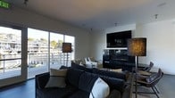 This contemporary single-floor space is designed with clean lines and a modern aesthetic that creates a sophisticated yet comfortable atmosphere. The living area serves as the centerpiece of the space, anchored by a sleek, wall-mounted flat-screen television above a linear gas fireplace with a glossy black surround — a striking focal point that adds warmth and visual interest to the room. Generous seating is provided by a large, plush sectional sofa in deep charcoal tones, accented with cream and neutral-toned throw pillows, complemented by additional lounge chairs for flexible arrangement. Stylish textured floor lamps flank the seating area, contributing to a warm and inviting ambiance. Large floor-to-ceiling sliding glass doors flood the space with abundant natural light and offer seamless access to an outdoor deck, where a railing-lined balcony reveals expansive waterfront views of the surrounding marina or waterway. The exterior scenery, visible through the glass, brings a sense of openness and connection to the outdoors. The interior palette is clean and neutral, with white walls and smooth ceilings that enhance the bright, airy feel of the space. Overall, this space strikes a balance between modern design and relaxed livability.