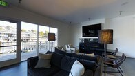 This contemporary single-floor space is designed with clean lines and a modern aesthetic that creates a sophisticated yet comfortable atmosphere. The living area serves as the centerpiece of the space, anchored by a sleek, wall-mounted flat-screen television above a linear gas fireplace with a glossy black surround — a striking focal point that adds warmth and visual interest to the room. Generous seating is provided by a large, plush sectional sofa in deep charcoal tones, accented with cream and neutral-toned throw pillows, complemented by additional lounge chairs for flexible arrangement. Stylish textured floor lamps flank the seating area, contributing to a warm and inviting ambiance. Large floor-to-ceiling sliding glass doors flood the space with abundant natural light and offer seamless access to an outdoor deck, where a railing-lined balcony reveals expansive waterfront views of the surrounding marina or waterway. The exterior scenery, visible through the glass, brings a sense of openness and connection to the outdoors. The interior palette is clean and neutral, with white walls and smooth ceilings that enhance the bright, airy feel of the space. Overall, this space strikes a balance between modern design and relaxed livability.
