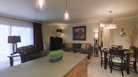 This single-floor residence offers an open-concept layout that seamlessly connects the kitchen, dining, and living areas into one cohesive and functional space. The kitchen features a generous countertop with a smooth matte finish and warm wood cabinetry below, complemented by contemporary pendant lighting that adds both task illumination and visual interest. The open flow leads directly into a comfortable living room furnished with a large sectional sofa accented with decorative throw pillows in earth tones. A framed landscape artwork anchors the main wall, while a tall floor lamp and a sleek black media console contribute to the room's balanced, layered aesthetic. Crown molding along the ceiling adds a refined architectural detail throughout. The dining area, visible to the right, includes bar-height seating and additional pendant lighting, creating a defined yet connected space for meals. Warm neutral wall tones, rich dark window drapery, and carpeted flooring unify the overall ambiance, giving the space a welcoming and polished feel. Natural light filters in through the windows, enhancing the warm, inviting atmosphere of this thoughtfully designed single-level home.