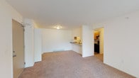 Located at 5001 E Riverside Dr in Evansville, Indiana, this single-floor unit offers a functional and well-organized layout suited for comfortable everyday living. The open living area is generously sized, featuring neutral-toned carpet and a clean, bright aesthetic enhanced by white walls and ceiling-mounted lighting. A half-wall partition connects the living space to the kitchen, which also features a pass-through countertop with a sink — ideal for casual dining or entertaining. The galley-style kitchen is efficiently designed with warm wood cabinetry, granite-patterned countertops, a stainless steel sink, and black appliances including a full-size refrigerator. Durable wood-look flooring grounds the kitchen space. The bedrooms are carpeted and well-lit, each benefiting from windows that bring in natural light and offer views of the surrounding greenery. Closets with built-in shelving and hanging rods provide practical storage. A wall-mounted air conditioning unit and baseboard heating are present throughout, ensuring year-round climate comfort. The layout, as seen in the dollhouse view, reflects a straightforward and efficient floor plan that maximizes usable space across the living, kitchen, and bedroom areas.