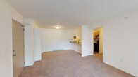 Located at 5001 E Riverside Dr in Evansville, Indiana, this single-floor unit offers a functional and well-organized layout suited for comfortable everyday living. The open living area is generously sized, featuring neutral-toned carpet and a clean, bright aesthetic enhanced by white walls and ceiling-mounted lighting. A half-wall partition connects the living space to the kitchen, which also features a pass-through countertop with a sink — ideal for casual dining or entertaining. The galley-style kitchen is efficiently designed with warm wood cabinetry, granite-patterned countertops, a stainless steel sink, and black appliances including a full-size refrigerator. Durable wood-look flooring grounds the kitchen space. The bedrooms are carpeted and well-lit, each benefiting from windows that bring in natural light and offer views of the surrounding greenery. Closets with built-in shelving and hanging rods provide practical storage. A wall-mounted air conditioning unit and baseboard heating are present throughout, ensuring year-round climate comfort. The layout, as seen in the dollhouse view, reflects a straightforward and efficient floor plan that maximizes usable space across the living, kitchen, and bedroom areas.