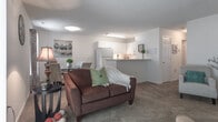 Located at 601 Hillpoint Blvd in Suffolk, Virginia, this single-floor residence offers a thoughtfully laid-out living space designed for comfort and functionality. The open living area is carpeted and filled with natural light streaming through large sliding glass doors fitted with vertical blinds, providing direct views of the surrounding greenery. The living space flows seamlessly into a dining area, creating a cohesive and inviting atmosphere.

The kitchen features white cabinetry with raised panel doors, light countertops, and a full suite of appliances including an electric range and over-the-range microwave, all set over rich hardwood-style flooring. A breakfast bar connects the kitchen to the adjacent living and dining areas.

The primary bedroom is generously sized with plush carpeting, a large closet, and a window dressed with vibrant curtains that add a pop of color. A second bedroom offers similar carpeted comfort, its own closet, and a serene, neutral palette. Both bathrooms feature hardwood-style flooring, white vanities with ample counter space, and tub-shower combinations. The entry foyer provides a welcoming transition into the home. Textured ceilings are consistent throughout, adding a classic residential character to the space.