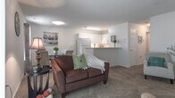 Located at 601 Hillpoint Blvd in Suffolk, Virginia, this single-floor residence offers a thoughtfully laid-out living space designed for comfort and functionality. The open living area is carpeted and filled with natural light streaming through large sliding glass doors fitted with vertical blinds, providing direct views of the surrounding greenery. The living space flows seamlessly into a dining area, creating a cohesive and inviting atmosphere.

The kitchen features white cabinetry with raised panel doors, light countertops, and a full suite of appliances including an electric range and over-the-range microwave, all set over rich hardwood-style flooring. A breakfast bar connects the kitchen to the adjacent living and dining areas.

The primary bedroom is generously sized with plush carpeting, a large closet, and a window dressed with vibrant curtains that add a pop of color. A second bedroom offers similar carpeted comfort, its own closet, and a serene, neutral palette. Both bathrooms feature hardwood-style flooring, white vanities with ample counter space, and tub-shower combinations. The entry foyer provides a welcoming transition into the home. Textured ceilings are consistent throughout, adding a classic residential character to the space.