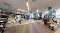 This contemporary fitness center offers a comprehensive, multi-zone workout environment designed to accommodate a wide range of exercise needs. The main workout floor is generously proportioned, with high ceilings and sleek linear pendant lighting that create a bright, energizing atmosphere. Large floor-to-ceiling windows along one wall flood the space with natural light and provide views of the surrounding greenery, enhancing the overall workout experience. The main floor is outfitted with an extensive array of cardio equipment, including treadmills, stair climbers, and stationary bikes, alongside a robust selection of strength training machines, cable systems, free weights, and benches. Structural columns punctuate the open floor plan, adding an architectural element to the space. A dedicated cycling studio features a row of stationary bikes, a large wall-mounted display screen, and full-wall mirror paneling, creating an immersive spin class environment. An adjoining movement studio offers light wood flooring, curved floor-to-ceiling mirrors, stability balls, medicine balls, and a Reflect interactive fitness mirror, ideal for guided workouts and yoga or stretching sessions. Entry to the facility is through double glass-paned doors, with dumbbell racks and accessory storage conveniently positioned near the entrance.