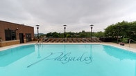 The Palisades presents an impressive rooftop amenity level designed for relaxation and leisure. The centerpiece of this outdoor space is a generously sized swimming pool with clear, turquoise water that reflects the surrounding environment. The pool deck is finished with warm-toned pavers and lined with a row of sleek, dark lounge chairs, providing ample space for sunbathing and unwinding. Tall, stylish pole-mounted light fixtures are evenly spaced along the deck, ensuring the space remains welcoming and well-lit into the evening hours. The elevated rooftop position affords expansive views of a lush, tree-lined horizon, creating a sense of seclusion and natural ambiance despite the urban setting. The open-air design allows for abundant natural light and fresh air, enhancing the resort-like atmosphere. Additional seating and accent planters frame the perimeter of the deck, adding architectural definition to the space. Whether used for a quiet morning swim or a social afternoon gathering, the rooftop pool area at The Palisades delivers a refined and inviting outdoor experience that stands as a signature feature of the property.