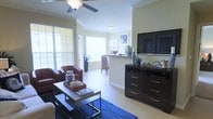 This single-floor space offers an open-concept layout that connects the living and dining areas in a cohesive, functional design. The living area features light hardwood-style flooring that extends throughout, lending a warm and airy feel to the interior. Crown molding lines the ceiling, adding a refined architectural detail, while a ceiling fan with a built-in light fixture provides both comfort and overhead illumination.

Large windows dressed with horizontal blinds line the walls, allowing abundant natural light to fill the space and creating a bright, welcoming atmosphere. The living area is anchored by a bold blue area rug and accommodates comfortable seating alongside a dark espresso-finished media dresser, which supports a large flat-screen television. Decorative accents, including coastal-inspired objects and greenery, contribute to the space's relaxed aesthetic.

Toward the rear, a partial wall with a granite-topped counter defines the transition to the dining area, where natural light continues to pour in through additional windows. A tall potted plant adds a natural element near the dining space. The overall ambiance is casual yet polished, combining functional layout with tasteful, coordinated furnishings.