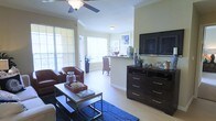 This single-floor space offers an open-concept layout that connects the living and dining areas in a cohesive, functional design. The living area features light hardwood-style flooring that extends throughout, lending a warm and airy feel to the interior. Crown molding lines the ceiling, adding a refined architectural detail, while a ceiling fan with a built-in light fixture provides both comfort and overhead illumination.

Large windows dressed with horizontal blinds line the walls, allowing abundant natural light to fill the space and creating a bright, welcoming atmosphere. The living area is anchored by a bold blue area rug and accommodates comfortable seating alongside a dark espresso-finished media dresser, which supports a large flat-screen television. Decorative accents, including coastal-inspired objects and greenery, contribute to the space's relaxed aesthetic.

Toward the rear, a partial wall with a granite-topped counter defines the transition to the dining area, where natural light continues to pour in through additional windows. A tall potted plant adds a natural element near the dining space. The overall ambiance is casual yet polished, combining functional layout with tasteful, coordinated furnishings.