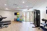 Located at 2855 W Commercial Blvd in Fort Lauderdale, Florida, this fitness facility is thoughtfully organized across two connected rooms, offering a complete workout environment within a compact, efficiently designed footprint. The space features light wood-look flooring throughout, white walls, and a drop ceiling with recessed lighting that creates a clean, bright atmosphere conducive to exercise.

The primary workout room is anchored by a wall-mounted flat-screen TV and benefits from generous natural light streaming through a set of elegant French doors topped with an arched transom window. These doors open to an outdoor green space, lending the room an open, airy feel. This area houses cardio equipment including treadmills, an elliptical, and a stationary bike, alongside a multi-functional cable machine, a rack of free weights with a workout bench, and an assortment of exercise balls, medicine balls, resistance bands, and fitness accessories organized on rolling carts.

The secondary room provides a quieter strength and recovery zone, featuring additional cardio machines, a cable machine, and a dumbbell rack. A standout element is the built-in wet bar area with dark espresso cabinetry, granite countertops, a subway tile backsplash, and a utility sink — ideal for post-workout convenience. A wall-mounted TV completes this functional space.