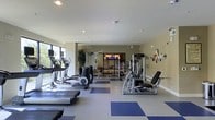 This single-floor fitness center offers a thoughtfully organized workout environment with a generous open layout. The space is finished with a durable, easy-to-clean floor featuring a two-tone grey and blue tile pattern that helps define distinct workout zones. Recessed lighting in the smooth white ceiling provides consistent, even illumination throughout the room, complemented by natural light streaming in through large windows along one wall, offering views of greenery outside.

The cardio section is well-stocked with treadmills, elliptical trainers, and other aerobic machines positioned to take advantage of the natural light. The strength-training area features multi-use cable and resistance machines, along with a dedicated free-weight and bench press station. A large mirror spans one wall in the strength zone, enhancing the sense of space and allowing users to monitor their form. A wall-mounted television is visible in the background, providing entertainment during workouts. Decorative wall art and potted plants add subtle aesthetic touches, contributing to a clean, modern ambiance throughout the facility.