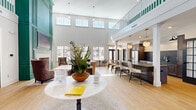 Located at 100 Top Deck Way in Smithfield, Virginia, this generously sized amenity center offers a diverse range of community spaces thoughtfully designed for both relaxation and activity. The centerpiece is a dramatic, double-height clubroom flooded with natural light from an abundance of large windows and an elevated mezzanine-level balcony accented with striking green railings. Light hardwood flooring, white columns, and pendant lighting contribute to the bright, welcoming atmosphere. A fully appointed kitchen area anchors one side of the clubroom, featuring dark cabinetry, stainless steel appliances, a large island with seating, and globe pendant lights — ideal for community gatherings and events. A lounge area with plush seating arrangements provides a comfortable setting for socializing or unwinding. The fitness center is expansively equipped with a full array of strength machines, free weights, treadmills, ellipticals, and other cardio equipment, all set on durable rubberized flooring beneath large ceiling fans and mirrored walls. A dedicated screening room features rows of leather reclining seats, framed movie posters, and a large-format display wall, delivering a true cinematic experience. The game room rounds out the amenities with a billiards table and casual seating. A connecting hallway ties all areas together seamlessly.