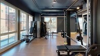 Located at 289 Huger St in Charleston, South Carolina, this single-floor fitness facility offers a thoughtfully designed workout environment with distinct zones for various training needs. The space features a cohesive industrial-modern aesthetic, characterized by deep navy and slate-blue walls, exposed ductwork along dark ceilings, and warm hardwood-style flooring that runs throughout.

The cardio area is positioned along a generous bank of floor-to-ceiling windows, allowing abundant natural light and views of the surrounding streetscape. Treadmills, ellipticals, and stationary bikes are arranged to take full advantage of this natural light, with wall-mounted monitors providing additional entertainment options.

A dedicated strength training zone houses a comprehensive selection of cable machines, weight benches, a rack with plate-loaded barbells, and multi-functional resistance equipment. Natural light filters into this area through large windows, creating a bright and energizing workout environment.

A flexible studio space, separated from the main floor by sleek black-framed glass doors, features a full mirrored wall and open floor space suited for group classes, stretching, or functional training. A medicine ball and dumbbell rack lines one wall, supporting a range of workout styles. Track lighting throughout the facility ensures consistent, well-distributed illumination across all zones.