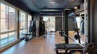 Located at 289 Huger St in Charleston, South Carolina, this single-floor fitness facility offers a thoughtfully designed workout environment with distinct zones for various training needs. The space features a cohesive industrial-modern aesthetic, characterized by deep navy and slate-blue walls, exposed ductwork along dark ceilings, and warm hardwood-style flooring that runs throughout.

The cardio area is positioned along a generous bank of floor-to-ceiling windows, allowing abundant natural light and views of the surrounding streetscape. Treadmills, ellipticals, and stationary bikes are arranged to take full advantage of this natural light, with wall-mounted monitors providing additional entertainment options.

A dedicated strength training zone houses a comprehensive selection of cable machines, weight benches, a rack with plate-loaded barbells, and multi-functional resistance equipment. Natural light filters into this area through large windows, creating a bright and energizing workout environment.

A flexible studio space, separated from the main floor by sleek black-framed glass doors, features a full mirrored wall and open floor space suited for group classes, stretching, or functional training. A medicine ball and dumbbell rack lines one wall, supporting a range of workout styles. Track lighting throughout the facility ensures consistent, well-distributed illumination across all zones.