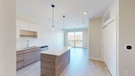 The Woodburn unit in Auburn Hills, Michigan offers a thoughtfully designed one-bedroom, one-bathroom layout that maximizes its efficient footprint. The open-concept living and kitchen area is anchored by light hardwood-style flooring and soaring ceilings that lend an airy, spacious feel. The contemporary kitchen is a standout, featuring stainless steel appliances including a French door refrigerator, electric range, microwave, and dishwasher. Sleek two-tone cabinetry—crisp white uppers paired with warm wood-grain lowers—complements the white quartz countertops and clean subway tile backsplash. A central kitchen island with pendant lighting serves as both a prep space and casual gathering point. The living area flows naturally toward a sliding glass door that opens to a private balcony, flooding the space with natural light and outdoor views. The bedroom is a comfortable retreat, carpeted for warmth and featuring a large window that brings in abundant daylight. A dedicated closet provides convenient storage. The bathroom is finished with a glass-enclosed walk-in shower with built-in shelving, a rainfall showerhead, and clean white surrounds. The unit also includes in-unit storage and a polished, move-in-ready aesthetic throughout.