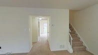 This two-story residence offers a straightforward and functional layout with a clean, neutral aesthetic throughout. The interior is finished with wall-to-wall carpeting that runs through the main living areas, complemented by crisp white walls that create a bright and airy atmosphere. A central hallway connects the main areas of the ground floor, leading toward a front entryway that allows natural light to filter into the space through a door with an exterior view of greenery beyond. The hallway is equipped with a ceiling-mounted light fixture and features a thermostat panel, highlighting the home's practical design. A floor-level ventilation register indicates a central HVAC system serving the space. The staircase, positioned to the side of the main living area, features carpeted treads and a white banister railing, providing a safe and accessible connection to the upper floor. The overall design is understated and neutral, offering a versatile canvas that can be adapted to a variety of interior styles. The home's layout emphasizes ease of movement between spaces, with open sightlines along the hallway and a well-defined separation between living areas and the vertical circulation to the second floor.