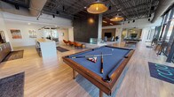 This expansive single-floor amenity space showcases a thoughtfully designed interior that blends industrial and contemporary aesthetics. The open-plan layout features exposed ductwork and structural ceilings in certain zones, contrasted by warm wood slat ceiling treatments in the lounge areas, creating distinct visual identities throughout the space.

At the heart of the main gathering area stands a dramatic dark brick fireplace column with a glass-encased hearth, serving as a striking architectural focal point. Surrounding it, light hardwood flooring extends throughout, anchoring clusters of modern seating including swivel lounge chairs in warm cognac leather and neutral tones. A full-size pool table occupies a central position, adding a recreational element to the space.

Multiple seating zones are defined throughout, each featuring flat-screen televisions mounted against textured feature walls. Floor-to-ceiling glass walls and doors flood the interior with natural light while offering panoramic views of an outdoor pool and landscaped courtyard. A bistro-style seating nook positioned directly against the glazed facade provides an intimate vantage point of the greenery beyond.

The kitchen area includes rich walnut cabinetry, a gas cooktop, stainless steel range hood, and a distinctive teal glass tile backsplash. A long communal counter with bar seating completes this functional zone, making the space well-suited for both social gatherings and everyday use.