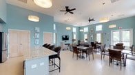 The Willows offers a welcoming single-floor clubhouse designed with both comfort and functionality in mind. The open-concept layout connects a bar counter area, casual dining space, and a relaxed lounge seamlessly, creating an inviting environment for socializing and gathering.

The interior is characterized by soft teal-painted walls that lend a calm, coastal ambiance throughout the space. Elevated ceilings with large windows along the walls allow abundant natural light to pour in, enhancing the airy and open feel. Ceiling fans are thoughtfully positioned throughout to maintain comfortable airflow.

The bar area features a generous granite countertop with wicker bar stools, ideal for casual conversation or a quick meal. Adjacent dining tables with matching wicker chairs provide additional seating options. The lounge area is furnished with plush sofas and accent chairs arranged around a wall-mounted flat-screen television, creating a cozy space for relaxation or entertainment.

Decorative pendant lighting adds warmth, complementing the light tile flooring that runs throughout. Framed botanical artwork on the walls ties the coastal aesthetic together. Overall, The Willows clubhouse presents a bright, tastefully designed communal space that balances relaxed comfort with a clean, modern style.