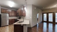 This single-floor residence offers a thoughtfully designed open-concept layout that seamlessly connects the kitchen and living areas. The kitchen is a standout feature, showcasing rich dark wood cabinetry that extends to the upper walls, providing ample storage. Granite countertops with subtle speckled detailing line the surfaces, including a peninsula with an integrated double sink and stylish pendant lighting hanging above — ideal for casual dining or meal prep. Stainless steel appliances, including a built-in microwave and gas range, complete the kitchen's functional appeal. The flooring throughout is a warm, dark hardwood that adds depth and character to the space. Vaulted ceilings in the kitchen area create an airy, open feel, while neutral gray walls provide a versatile backdrop. An arched doorway adds an architectural touch, transitioning gracefully into adjacent areas. Natural light filters in through multiple windows and a glass-paneled entry door, brightening the open living space. A staircase visible in the layout suggests additional levels or access to lower areas. Overall, the space balances modern practicality with classic design elements, making it both inviting and highly functional.