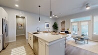 This thoughtfully designed single-floor residence showcases a clean, contemporary aesthetic throughout. The open-concept kitchen is a standout feature, outfitted with white cabinetry, stainless steel appliances, a subway tile backsplash, and a large central island with quartz countertops and pendant lighting overhead — seamlessly connecting to the living area. The living room is generously proportioned, featuring light wood-style flooring, a ceiling fan, built-in shelving, and large windows that invite natural light. A dedicated office or study space includes arched metal shelving units and a substantial work surface, offering a stylish and functional workspace. The bedrooms are carpeted for comfort, with the primary suite featuring a window with outdoor views, soft neutral tones, and direct access to a luxurious en-suite bathroom. The primary bath is expansive, with a double vanity, soaking tub, glass-enclosed shower, large-format tile flooring, and a walk-in closet. A secondary bathroom provides a clean, modern vanity with access to an additional walk-in closet. Soft gray walls and consistent finishes throughout create a cohesive and inviting atmosphere across every room.