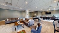 Located at 345 S Depeyster St in Kent, Ohio, this single-floor community lounge offers a generously sized, open-plan layout designed for both relaxation and entertainment. The space features warm hardwood-style flooring throughout, complemented by neutral gray-toned walls and a drop tile ceiling with recessed lighting that creates a bright, welcoming atmosphere.

The main lounge area is anchored by multiple plush brown leather sofas and armchairs arranged around low-profile coffee tables, providing comfortable seating for groups. A full-size pool table serves as a central entertainment feature, while a mounted flat-screen television and a branded accent wall add to the recreational appeal.

Toward the rear of the space, large windows with roller shades allow ample natural light to fill a dedicated dining area, furnished with high-top tables and bar-height chairs. Adjacent to this is a wet bar and counter space equipped with a coffee station and small appliances, ideal for casual gatherings.

A hallway connects the main area to additional sections of the building, and structural columns are thoughtfully integrated into the open floor plan without disrupting the flow. The overall design strikes a balance between modern aesthetics and functional comfort, making it a well-appointed communal space suited for socializing, studying, or unwinding.